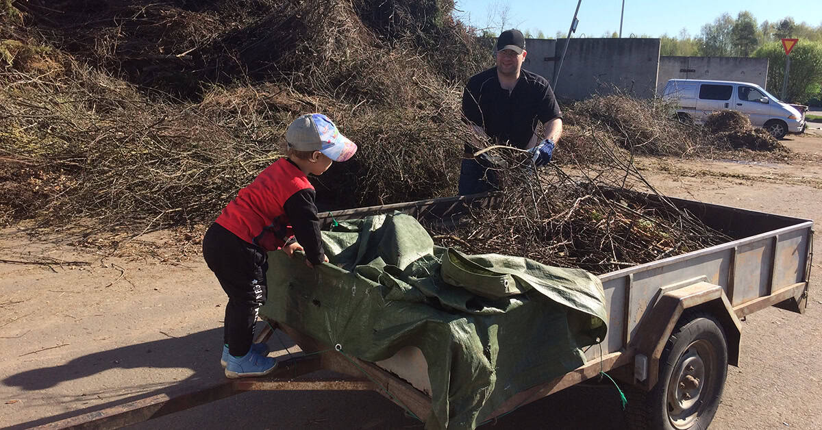 Lapsi tuo isänsä kanssa puutarhajätteitä peräkärrillä. Taustalla iso kasa puutarhajätettä.