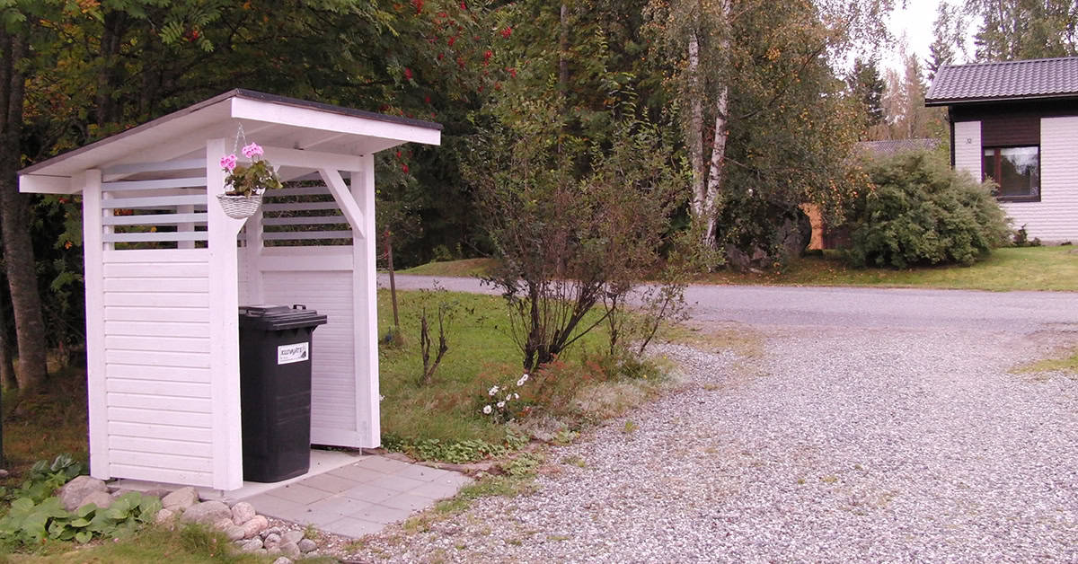 Näkymä omakotitalon pihatielle, vasemmassa laidassa valkoinen jätekatos, jonka räystäässä roikkuu kukka-amppeli.